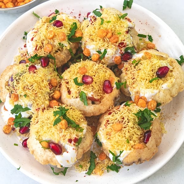 Crispy Indian chaat topped with yogurt, sev, pomegranate seeds, and fresh cilantro on a white plate.