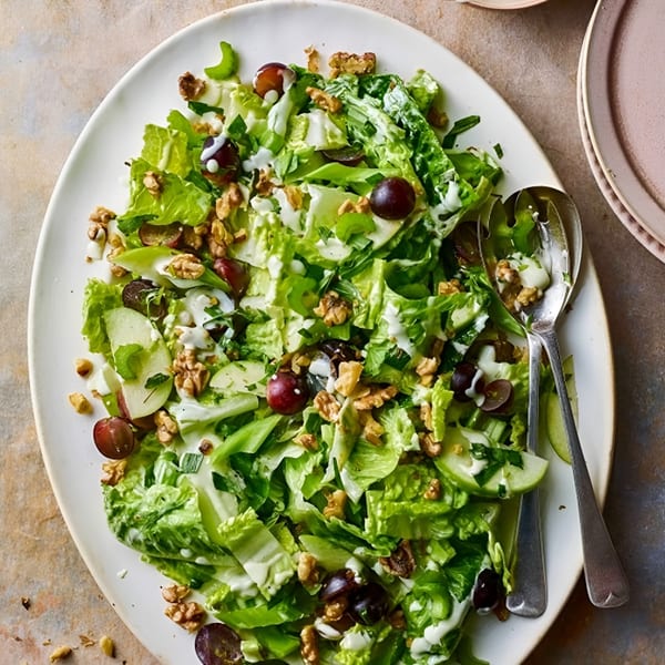 Fresh green salad with grapes, walnuts, and creamy dressing on a white platter with serving spoons.