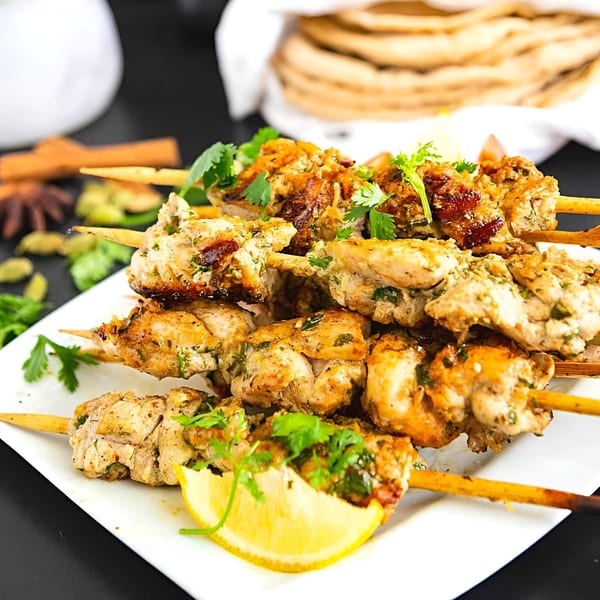 Plate of chicken skewers with fresh cilantro and lemon, alongside flatbread and spices.